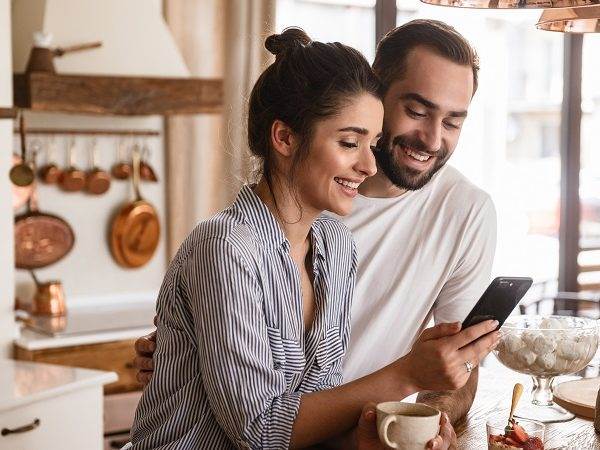Photo of smiling brunette couple drinking coffee and using cell phone during breakfast at home