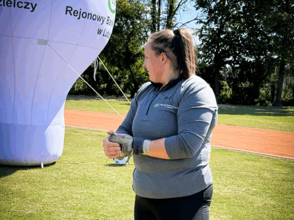 Kobieta-lekkoatletka, stojąca na trawie, trzyma ciężar do rzutu młotem. W tle widoczny jest duży balon reklamowy banku i drzewa. W dolnej części zdjęcia znajduje się logo 