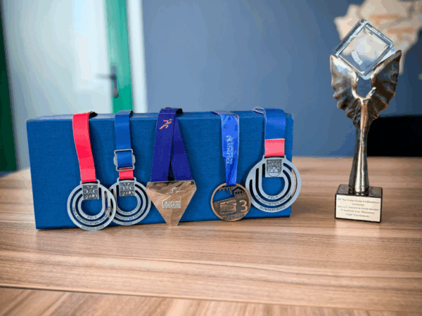 Na drewnianym biurku w biurze banku leżą medale sportowe i statuetka. W tle widoczna jest drewniana mapa oddziałów banku na ścianie. W dolnej części zdjęcia znajduje się logo 