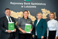 Cztery osoby, w tym przedstawiciele banku i lekkoatletka, stoją w biurze banku, prezentując zielone teczki. W tle widoczna jest duża mapa Polski na ścianie. W dolnej części zdjęcia znajduje się logo 
