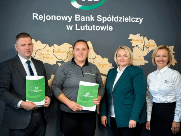Cztery osoby, w tym przedstawiciele banku i lekkoatletka, stoją w biurze banku, prezentując zielone teczki. W tle widoczna jest duża mapa Polski na ścianie. W dolnej części zdjęcia znajduje się logo 