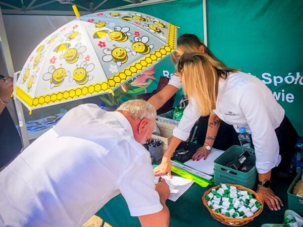 na zdjęciu nachylony nad stołem mężczyzna zasłaniający się parasolem w pszczółki, z prawej strony w tle stoją dwie pracownica Banku