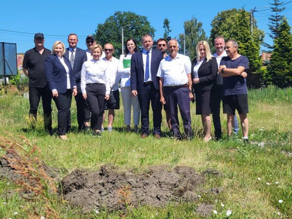 grupa osób, dziewięciu mężczyzn, cztery kobiety, uśmiechnięci, stoją na działce budowlanej,