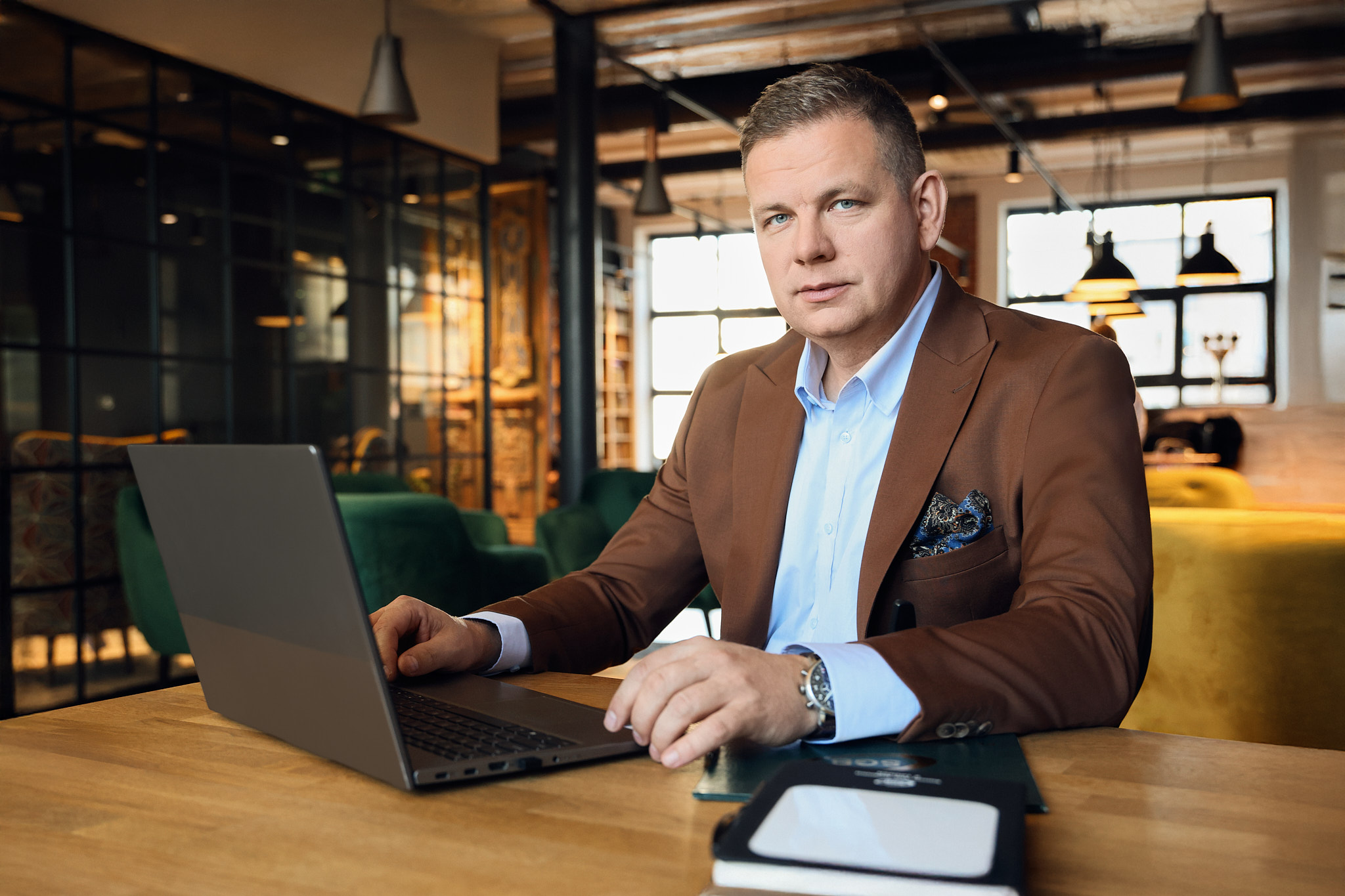 na zdjęciu mężczyzna siedzący przy biurku, na biurku laptop oraz notes, w tle pomieszczenia biurowe