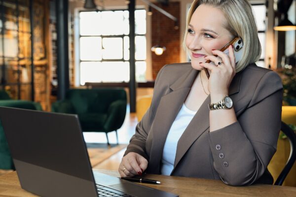 na zdjęciu kobieta siedząca przy biurko, w dłoni przy uchu trzyma telefon, na biurko, stoi laptop